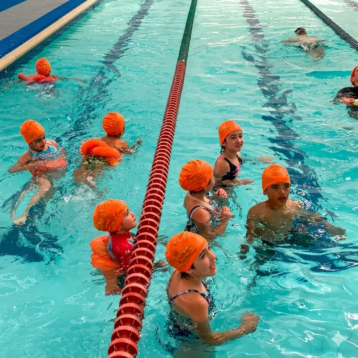 Niños con gorros naranjas practicando en una piscina, junto a un carril de natación.
